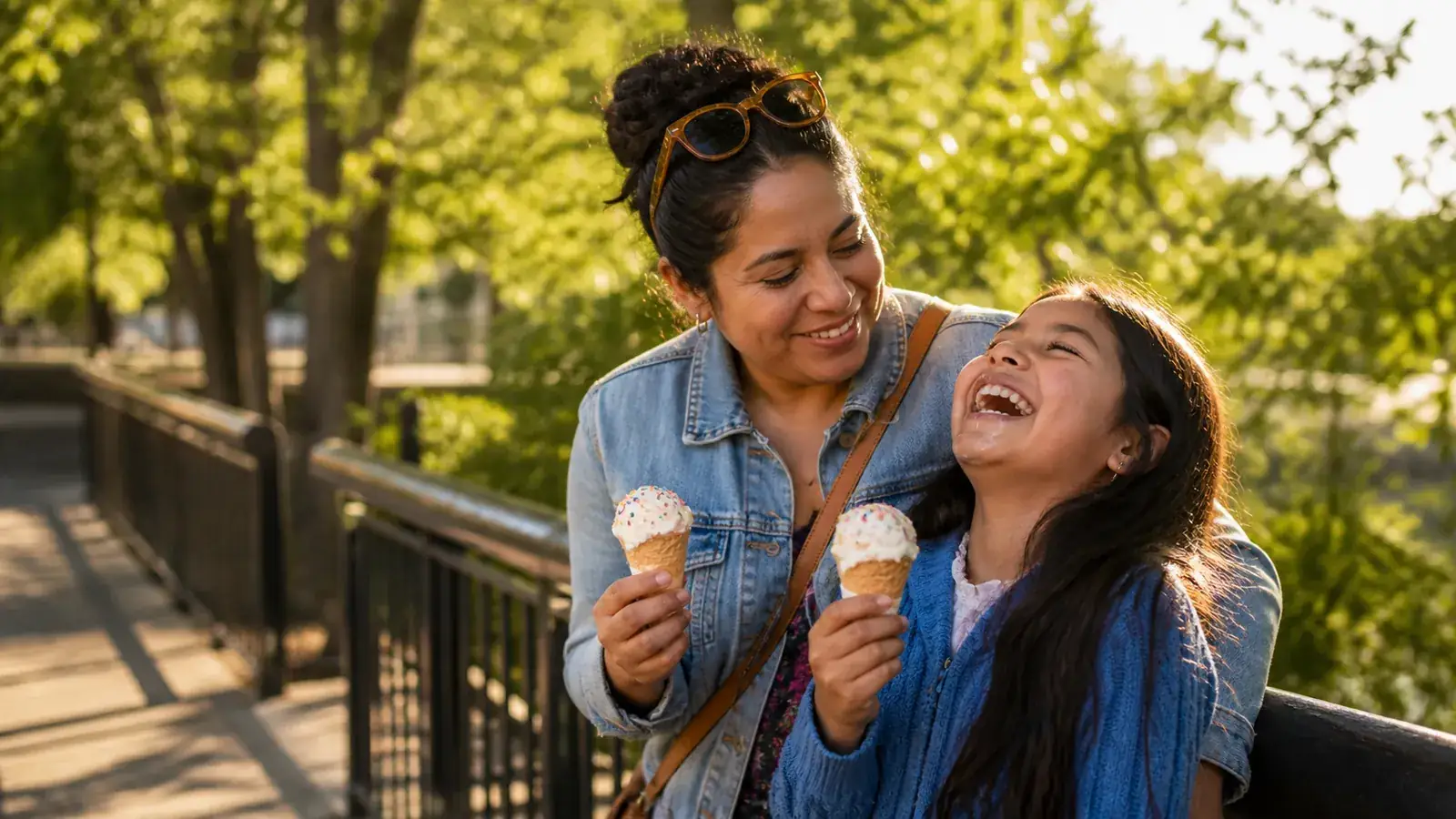 Mother and daughter enjoying ice-cream cones by a sunny park railing, both laughing